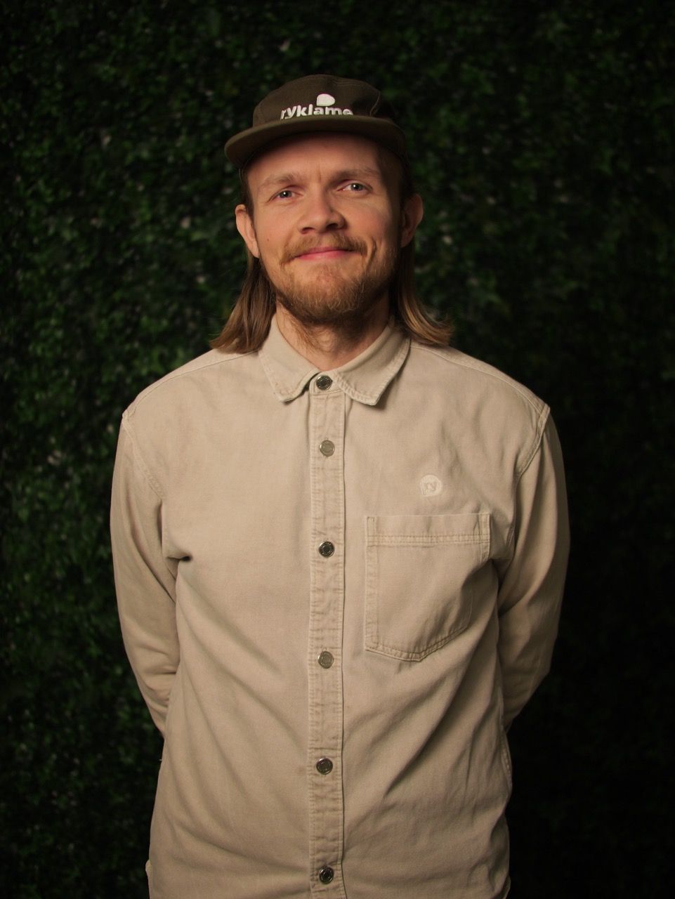 Tom Egill smiler i ryklame-caps foran en mørkegrønn løvvegg.